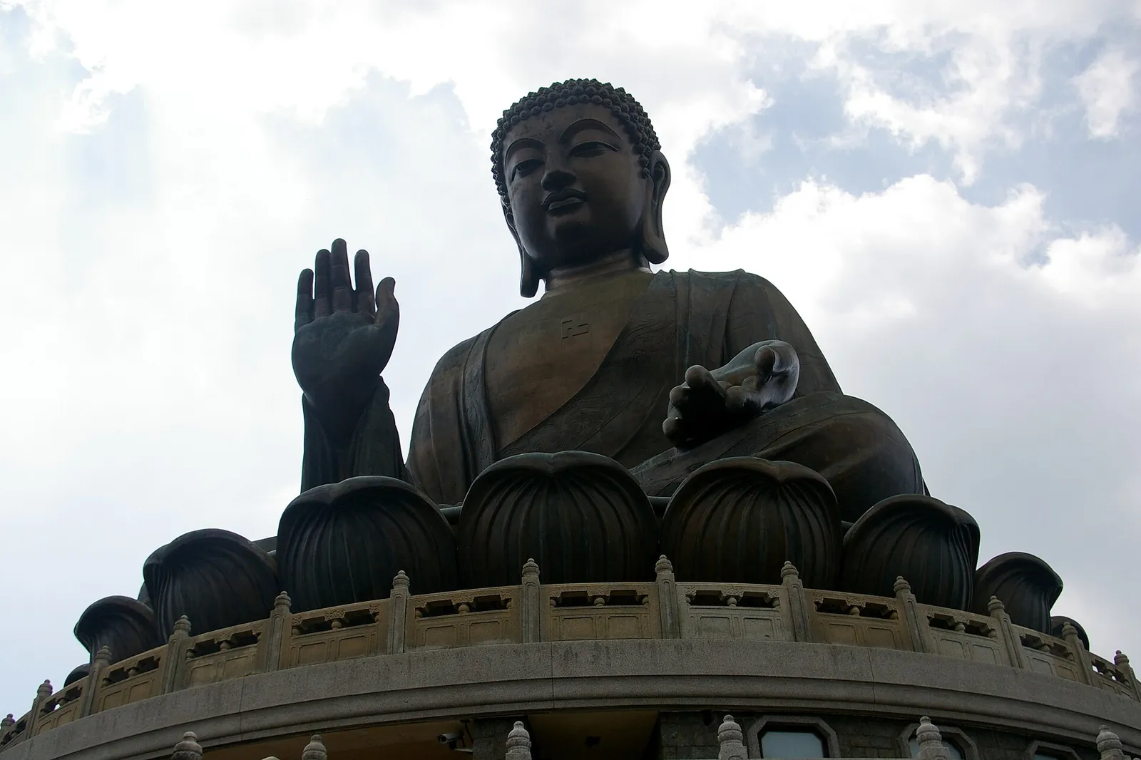 Bronzový Tian Tan Buddha viděný zespodu, s jednou zdviženou rukou, usazený na lotosovém trůnu proti zamračené obloze