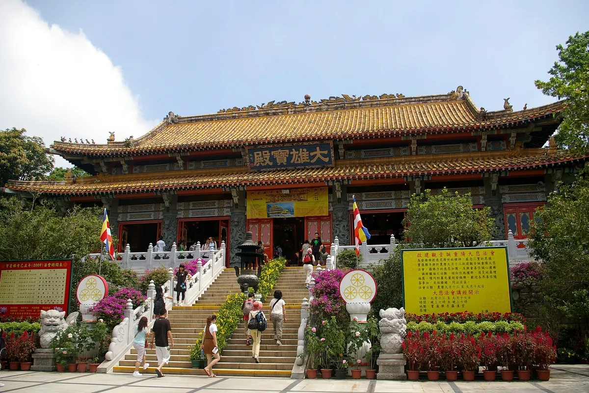 Hlavní síň Po Lin Monastery se žlutou taškovou střechou a červenými sloupy, k níž vede široké bílé schodiště s květinovými záhony po obou stranách