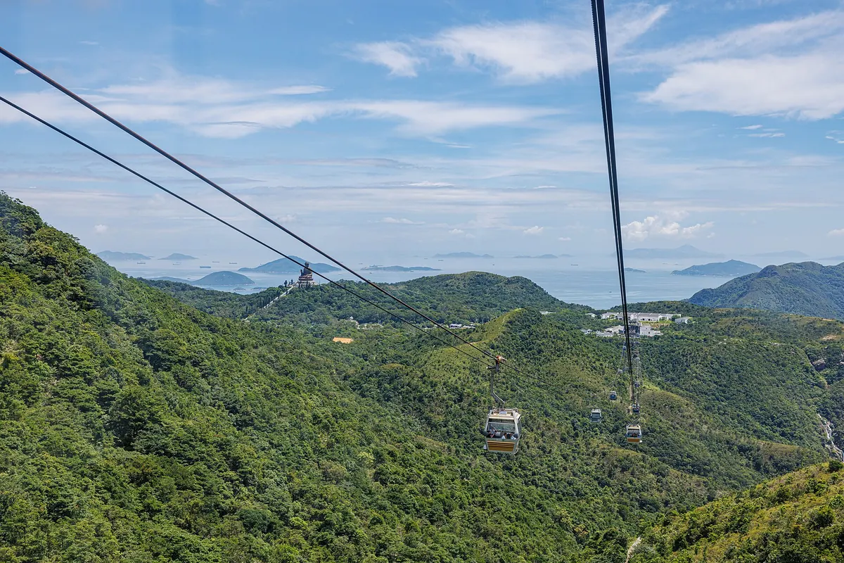 To kabler og en kabine, der krydser de skovklædte bjergkamme på Lantau, med Tian Tan Buddha på en fjern bjergtop og havet og øerne bagved