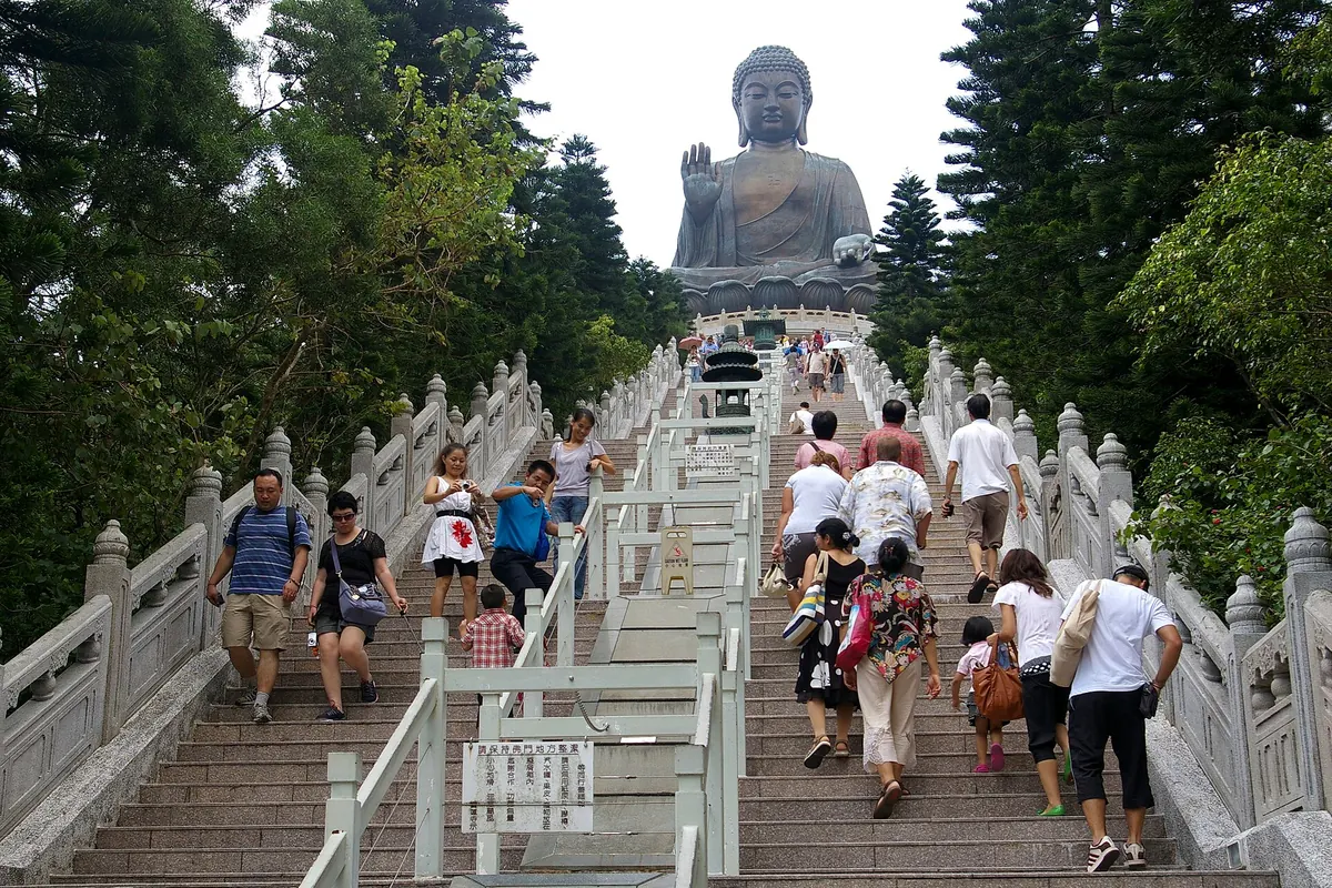 Široké kamenné schodiště s bílým zábradlím stoupající k bronzovému Tian Tan Buddha, s návštěvníky na schodech a stromy po obou stranách