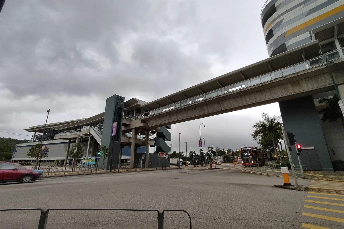 Das niedrige, geschwungene Dach des Seilbahnterminals Tung Chung neben einer Fußgängerbrücke und einer breiten Straße unter bewölktem Himmel